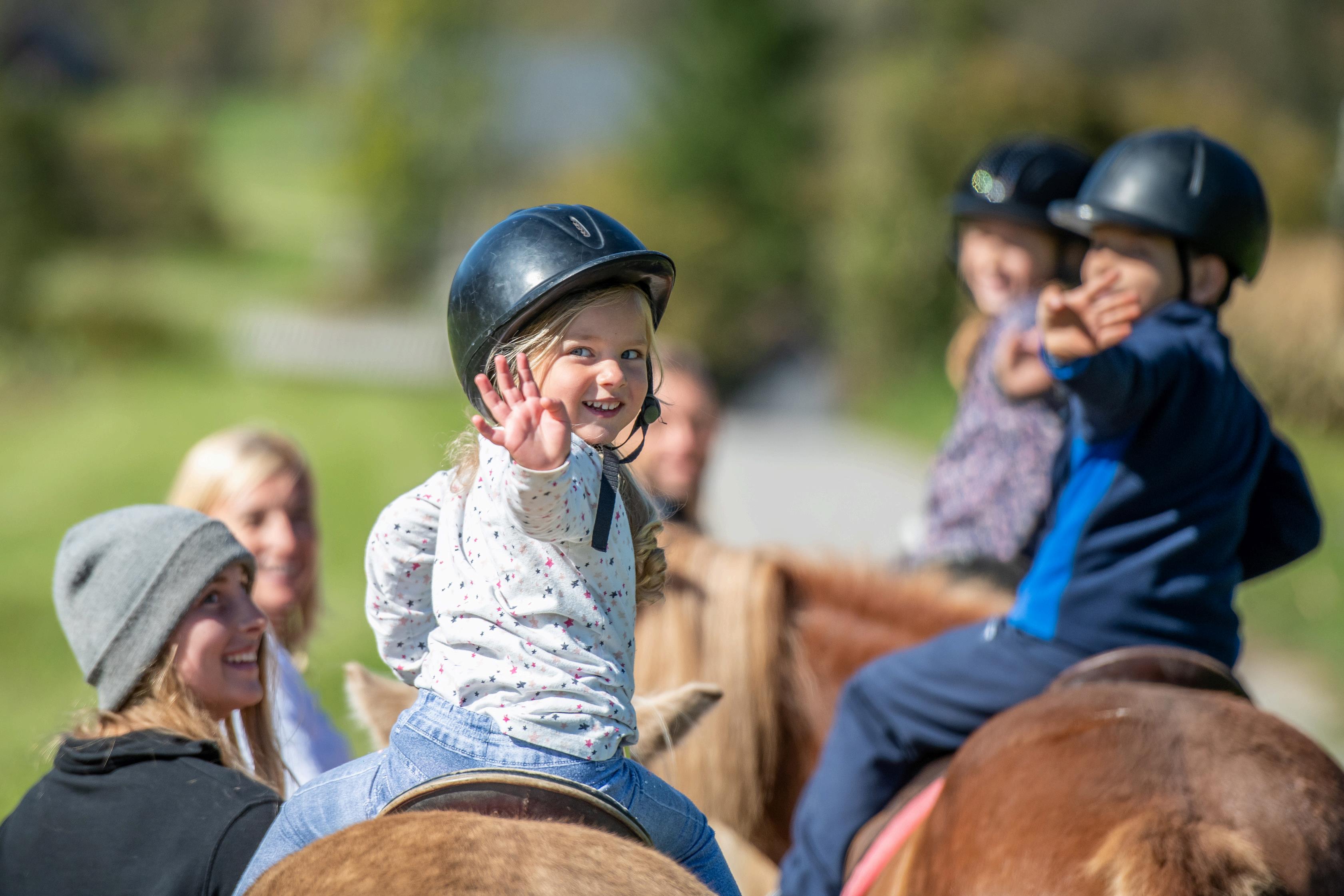 “A group of young children on horses with volunteers leading them away and the horses are turned back and waving at the camera